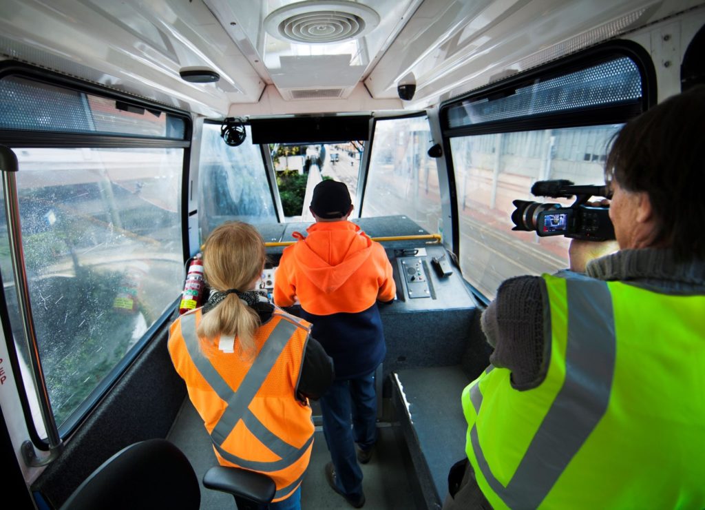 Three people are standing inside a Sydney Monorail driver's cab wearing high-viz vests. The front window of the monorail car is raised and the track ahead can be seen. The Monorail is travelling along the track and the view from the side windows is blurred as it travels along. A tall man is operating a video camera, the driver operates the monorail and a young woman stands behind him.